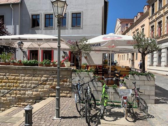 Auf einer Straße, stehen in der Mitte des Bildes Fahrräder, dahinter eine Mauer, auf der Mauer links und rechts kleine grüne Bäume, dazwischen ein Sonnenschirm, links neben den Fahrrädern ist eine dunkle Laterne, neben der Laterne sind Treppen, im Hintergrund sind mehrere Gebäude bei sonnigem Wetter.