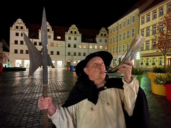 Auf dem Marktplatz, der Stadt Torgau, steht bei Nacht ein als Nachtwächter verkleideter Mann, mit Speer in der Hand und Horn im Mund, im Hintergrund das Gebäude des Rathauses