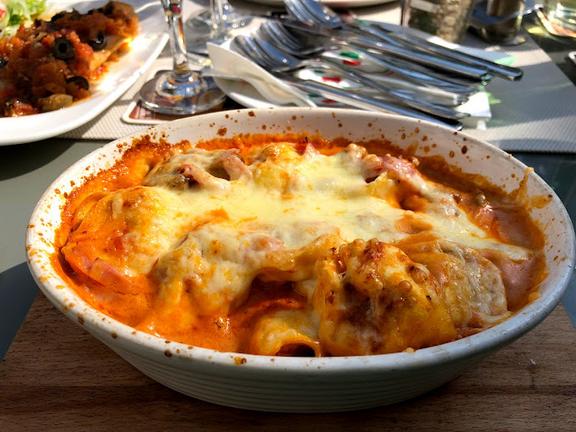 Auf einem braunen Holztisch, steht ein weißer tiefer Teller mit Lasagne, im Hintergrund silbernes Besteck und ein Teil eines anderen Gerichtes.