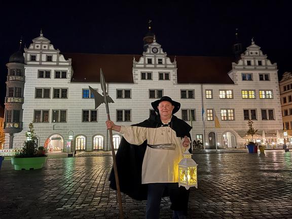Auf dem Markplatz, der Stadt Torgau, steht ein als Nachtwächter verkleidete Person bei Nacht, mit ausgestrecktem rechten Arm in welcher er einen Speer hält, in der linken Hand hält er eine Laterne hoch