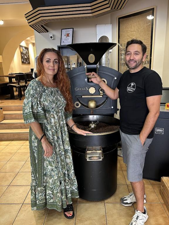 Zwei Personen stehen neben einer großen, schwarzen Kaffeeröstmaschine in einem modern eingerichteten Café oder Rösterei; die Frau trägt ein langes, gemustertes Kleid, der Mann ein schwarzes T-Shirt und graue Shorts.