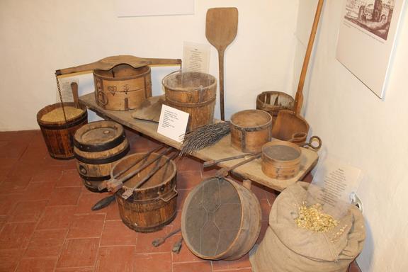 Verschiedene historische Brauwerkzeuge und Behälter aus Holz, darunter Fässer, Bottiche und ein Mehlsack mit Getreide, ausgestellt in einem Museumseck.