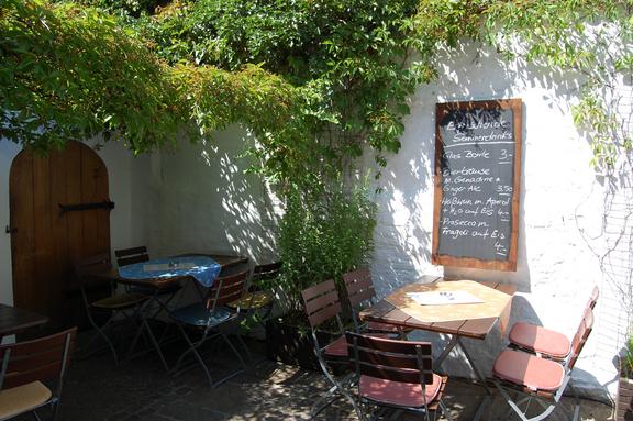 Außenansicht des Biergartens Herr Käthe, mit weißer Wand, Speisetafel, einer Sitzmöglichkeit und einer grünen Hecke.