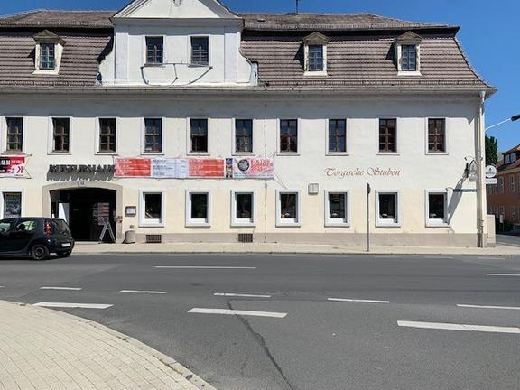 Ein großes, weißes Gebäude mit mehreren Fenstern und einem alten Dach. Vor dem Haus hängen Werbebanner, und links steht ein schwarzes Auto an der Straße.