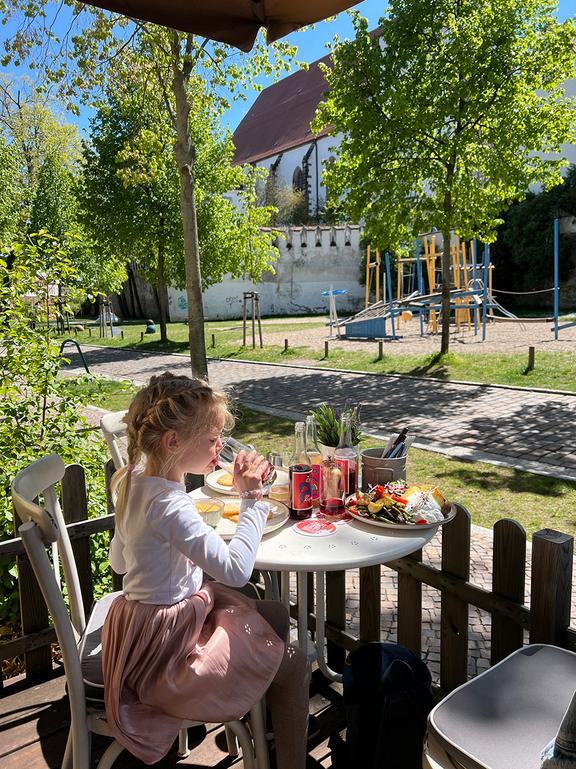 in einem Restaurant draußen auf einem Stuhl an einem Tisch vor einem Zaun sitzendes Mädchen mit essen und trinken auf dem Tisch bei sonnigem Wetter 