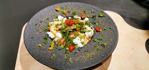 Eine grauer tiefer Teller, stehend auf dunklem und hellem Untergrund, in der Mitte eine helle Speise mit Kräutern und Tomaten. 