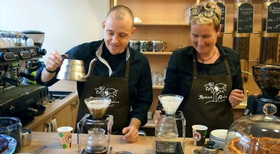Zwei Baristas in schwarzer Arbeitskleidung bereiten mit Handfiltermethoden Kaffee zu, umgeben von Kaffeebohnen, Tassen und professioneller Ausstattung in einem gemütlichen Café.