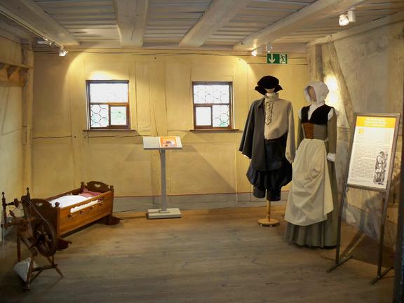 Historische Kleidung in einem Museum mit Fenstern und einer Krippe.
