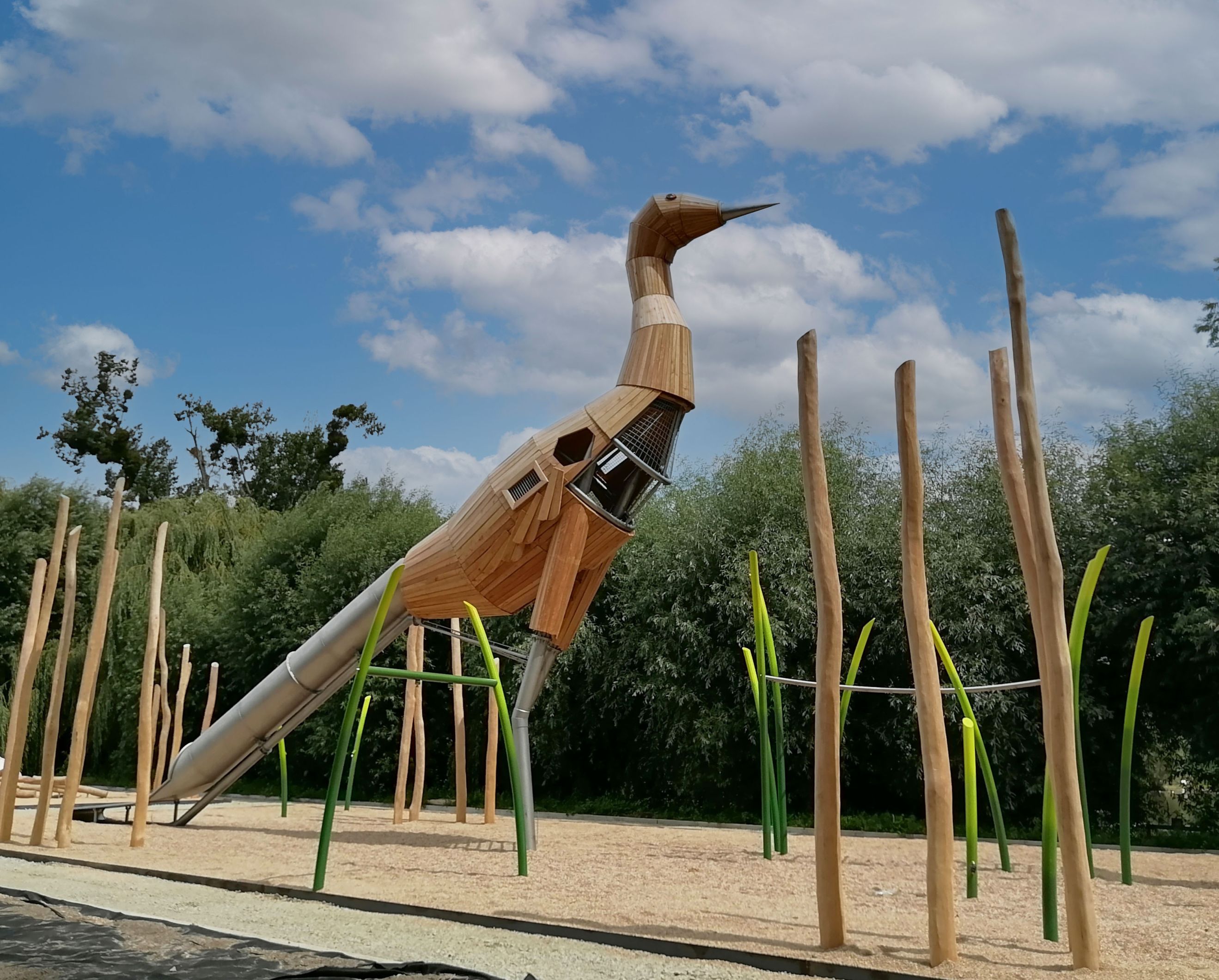 Große hölzerne Kranichskulptur auf einem Spielplatz mit Stangen und grünen Kletterelementen unter bewölktem Himmel