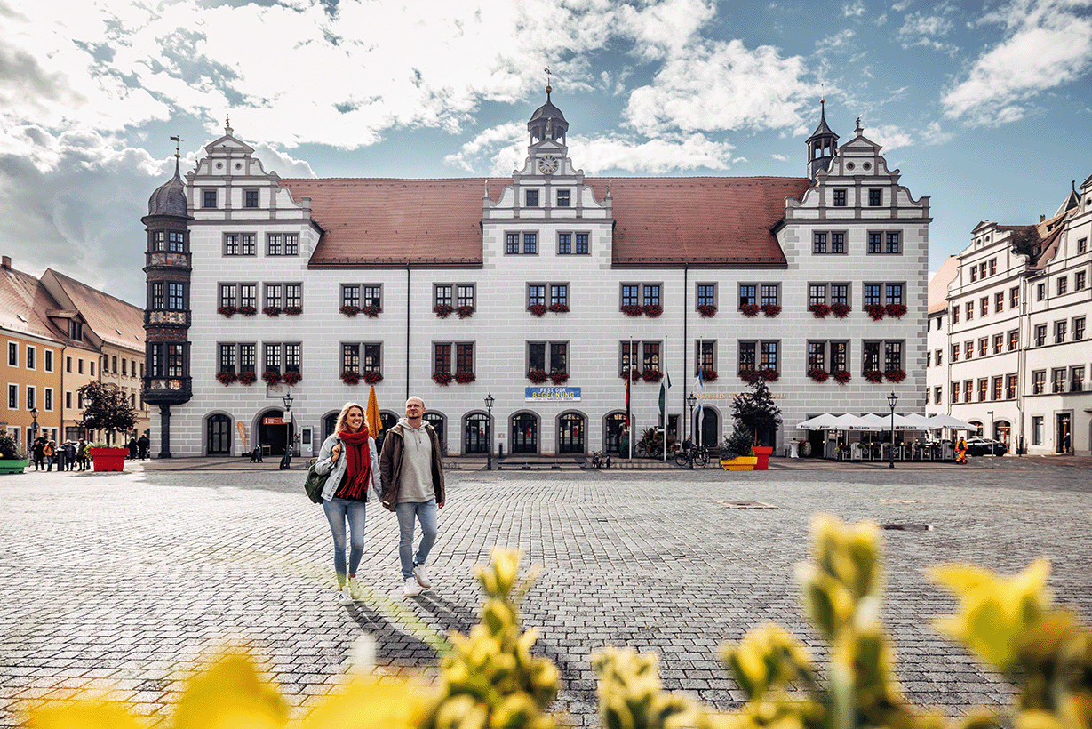 Marktplatz in Torgau mit dem historischen Renaissance-Rathaus und zwei Personen, die über den gepflasterten Platz gehen, bei bewölktem Himmel