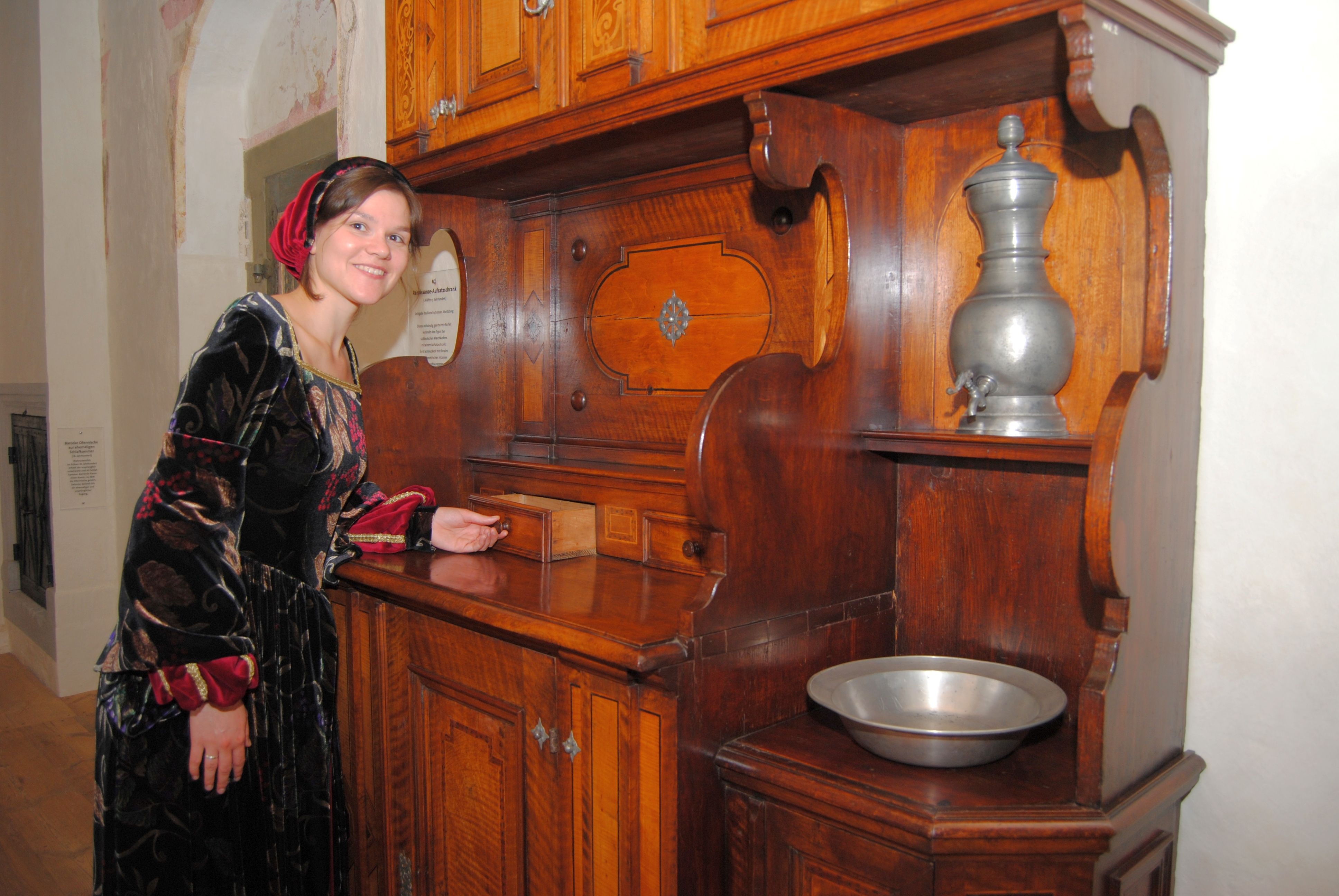 Eine Frau in historischer Kleidung steht neben einem antiken Küchenschrank.